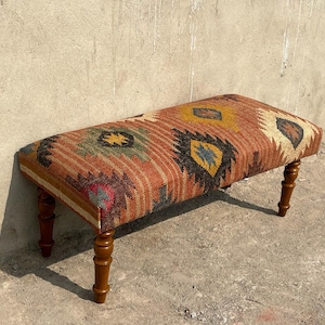 May include: A rectangular bench with a patterned textile top and four wooden legs. The textile features a mix of colors including orange, green, yellow, and black. The legs are a dark brown color.