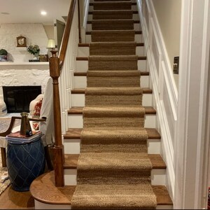 May include: A natural fiber stair runner in a light brown color is placed on a set of white painted wooden stairs. The runner is made of a woven material with a textured surface.