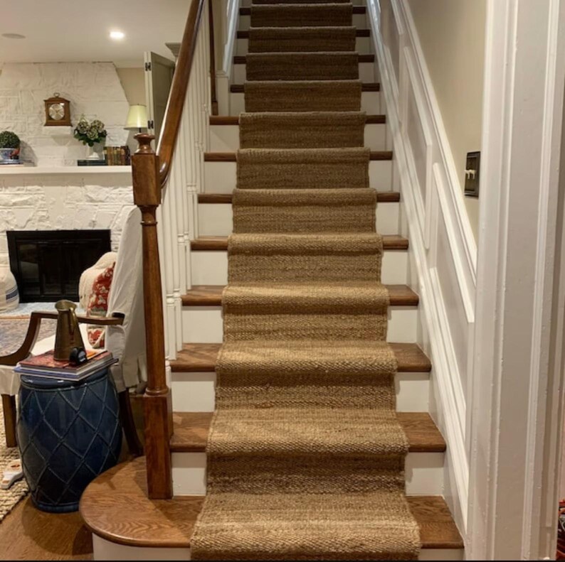 May include: A natural fiber stair runner in a light brown color is placed on a set of white painted wooden stairs. The runner is made of a woven material with a textured surface.