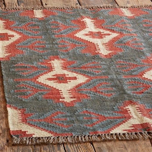 May include: A rectangular rug with a geometric pattern in shades of gray, red, and cream. The rug has a fringed edge and is displayed on a wooden surface. The design features diamond and arrow-like shapes.