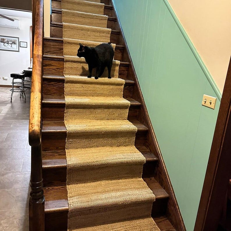 May include: A black cat walks up a set of wooden stairs with a natural fiber runner. The stairs have a dark wood finish and the wall is painted a light teal color.