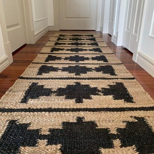 May include: A black and natural jute rug with a geometric pattern. The rug is laid out in a hallway with white walls and wooden floors.
