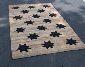 May include: A rectangular jute rug with a repeating pattern of black eight-pointed stars. The rug has a natural beige color with the dark stars evenly spaced across the surface. The rug is suitable for indoor or outdoor use.