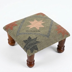 May include: A small, square footstool with a woven, patterned top. The top features a central star design in peach, surrounded by geometric shapes in red, gray, and black on a green background. The footstool has four short, turned wooden legs.