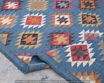 May include: A blue rug with a geometric pattern. The rug features repeating square designs in shades of orange, red, brown, and white. The rug is folded over, revealing the underside and the texture of the material.