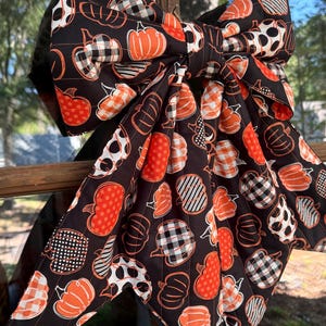 May include: A large decorative bow made of black fabric featuring a repeating pattern of orange pumpkins. The pumpkins are decorated with various white and black patterns, including polka dots, buffalo check, and thin stripes, with long fabric tails.
