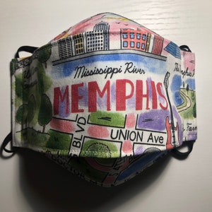 May include: A fabric face mask with a colorful Memphis, Tennessee design. The mask features the text "Memphis" in large red letters, along with illustrations of buildings, a guitar, and street names like Union Ave and Doctor M.L.K. Blvd.