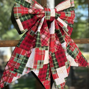May include: A large decorative bow made from fabric featuring a patchwork of red, green, and white plaid patterns. The bow has two prominent loops at the top and two long, pointed tails hanging down, with subtle glitter accents on the fabric surface.