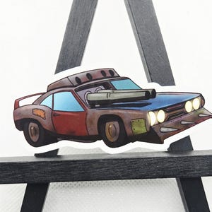 May include: A sticker featuring a cartoon illustration of a customized car. The car has a brown body with red and blue accents, large exhaust pipes, and a unique front design. The car appears to be a post-apocalyptic vehicle.