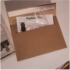 Puede incluir: Un sobre de papel marr&oacute;n con una ventana de pl&aacute;stico transparente. La ventana muestra un albar&aacute;n con el texto "Packing Slip" y "Order # 2425722457". Tambi&eacute;n hay una peque&ntilde;a pegatina con el texto "CitaDraws" y una imagen de dibujos animados de una persona.