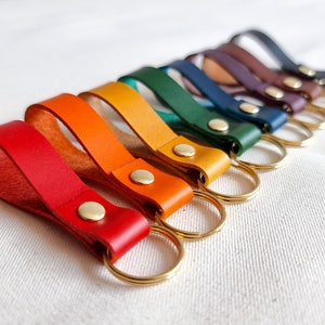 May include: A set of ten leather keychains in a rainbow of colors, each with a gold metal ring. The keychains are arranged in a row, with the red keychain at the bottom and the purple keychain at the top.