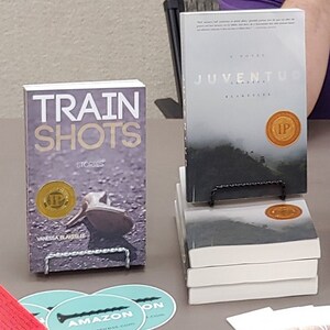 Może przedstawiać: Dwie książki wystawione na podpisywanie. Książka po lewej stronie nosi tytuł "Train Shots" z nazwiskiem autorki Vanessa Blakeslee. Książka po prawej stronie nosi tytuł "Juventud" z nazwiskiem autorki Vanessa Blakeslee. Obie książki mają złotą pieczęć z literami "IP" na nich.