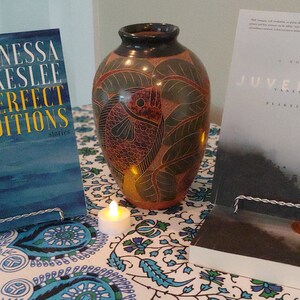 Może przedstawiać: Dwie książki Vanessy Blakeslee, "Perfect Conditions" i "Juvenitude", są wystawione na stole z brązowym ceramicznym wazonen z wzorem ryby. Stół ma biało-niebieski obrus w kwiaty i znajdują się na nim dwie białe świeczki.