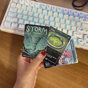 May include: Three rectangular trading cards featuring colorful illustrations. One card displays a green tornado with the text Storm Counter. Another card shows a green circular pool with the text Sol Ring. The third card depicts a red structure labeled Roaming Throne.
