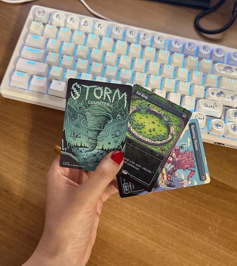 May include: Three rectangular trading cards featuring colorful illustrations. One card displays a green tornado with the text Storm Counter. Another card shows a green circular pool with the text Sol Ring. The third card depicts a red structure labeled Roaming Throne.