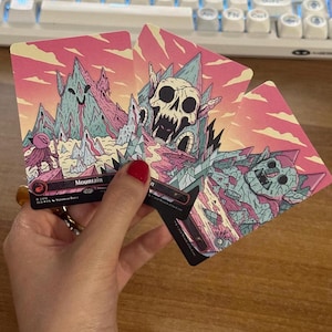 May include: Three rectangular trading cards featuring stylized mountain illustrations in shades of pink, teal, and cream. The cards display artistic depictions of rocky peaks, a large skull, and a stone creature, with the text Mountain printed on one.