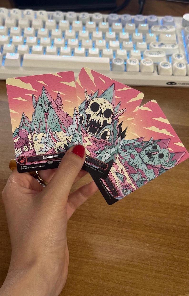 May include: Three rectangular trading cards featuring stylized mountain illustrations in shades of pink, teal, and cream. The cards display artistic depictions of rocky peaks, a large skull, and a stone creature, with the text Mountain printed on one.