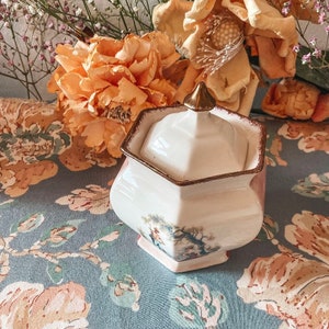 May include: A white porcelain sugar bowl with a gold trim and a floral design. The bowl has a lid with a gold knob. The bowl is sitting on a floral patterned fabric.