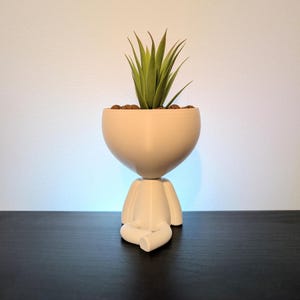 May include: A matte white 3D-printed planter shaped like a seated figure with crossed legs. The rounded head section serves as the container, holding a green succulent plant with pointed leaves and small brown pebbles on the surface of the soil.