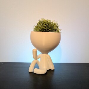 May include: A white 3D-printed planter shaped like a seated figure with one arm raised to its head, holding a small, spiky green air plant. The smooth, matte-finish container features a rounded bowl top and a minimalist, stylized human form.