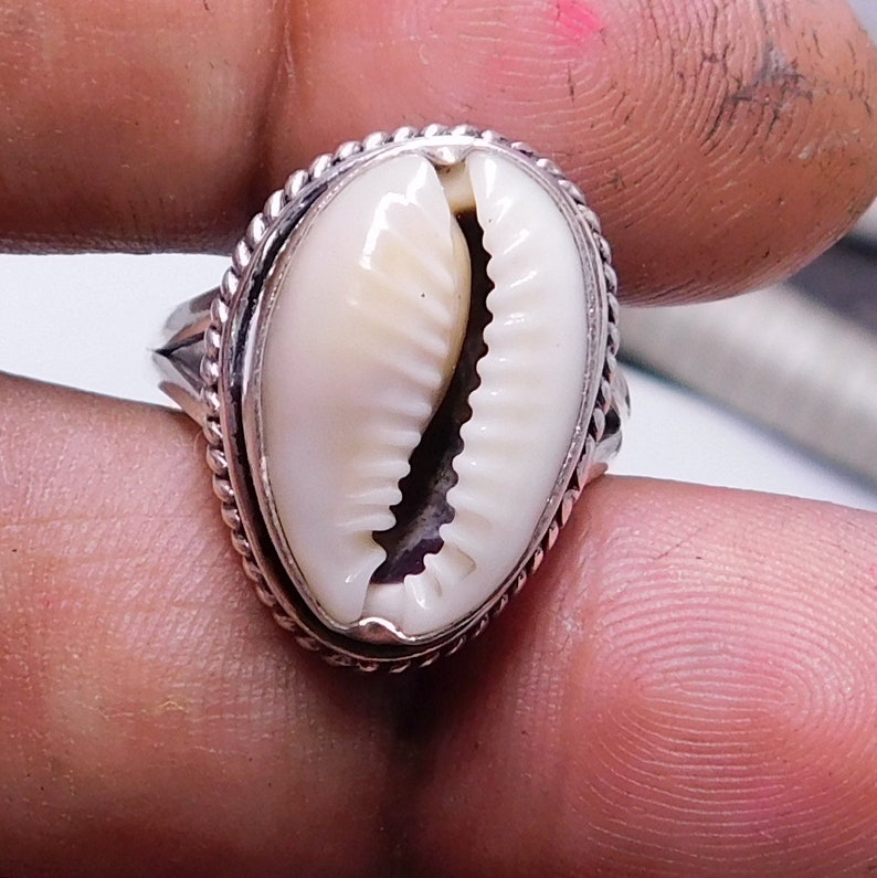 Op de afbeelding: Een zilveren ring met een witte kauri schelp in het midden. De ring heeft een decoratieve rand rond de schelp.