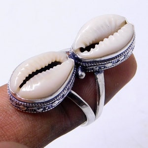 Op de afbeelding: Een zilveren ring met twee witte kauri-schelpen die in de band zijn gezet. De schelpen zijn naar elkaar gericht en hebben een gedetailleerd zilveren ontwerp rond de randen.