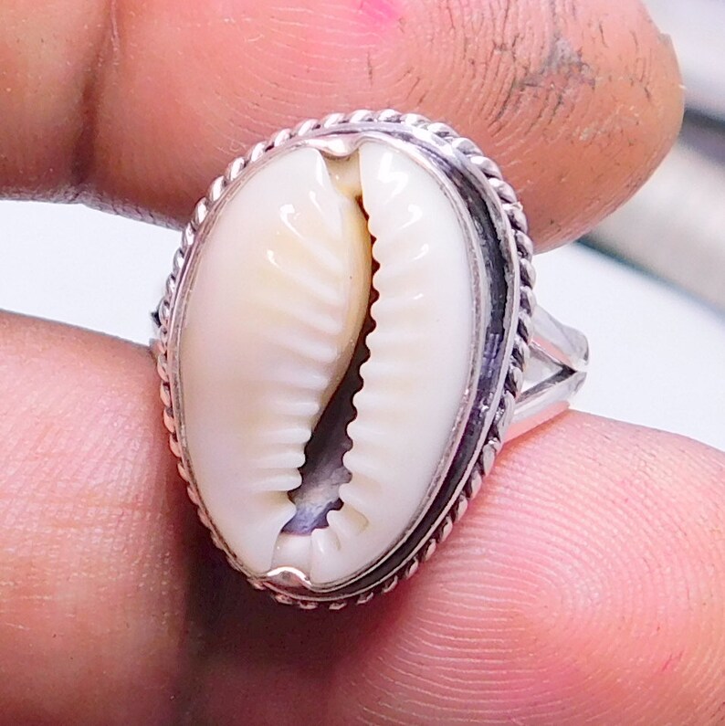 Op de afbeelding: Een zilveren ring met een witte kauri schelp in een touwachtige rand.