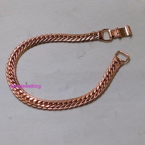 May include: A copper-coloured bracelet with a curb chain design. The bracelet features a rectangular clasp and a D-ring closure. The chain links are closely interlocked, creating a textured surface. The bracelet is displayed on a light-coloured surface.