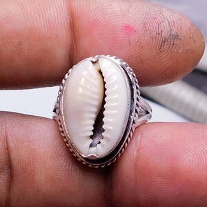 Op de afbeelding: Een zilveren ring met een witte kauri schelp in het midden. De ring heeft een gevlochten ontwerp rond de rand.