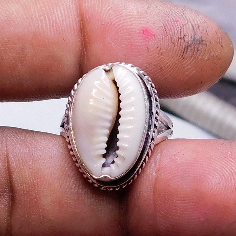 Op de afbeelding: Een zilveren ring met een witte kauri schelp in het midden. De ring heeft een gevlochten ontwerp rond de rand.