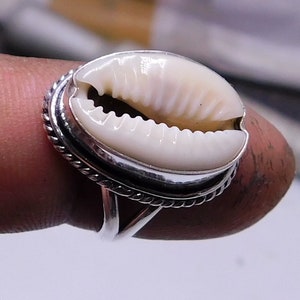 Op de afbeelding: Een zilveren ring met een witte kauri schelp in het midden. De ring heeft een touwachtig ontwerp rond de band.