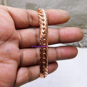 Peut inclure: Un bracelet de couleur cuivre poli présentant un design à maillons épais et entrelacés. La surface métallique possède une finition brillante et réfléchissante avec un motif tressé répétitif sur toute la longueur, affichant le texte silverjewelling.