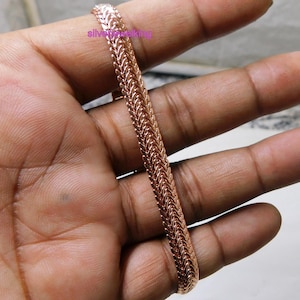Puede incluir: Una pulsera de oro rosa con un diseño tejido. La pulsera se sostiene en una mano, mostrando su intrincado patrón y cálido color metálico. Se destaca la textura y el brillo de la pulsera.