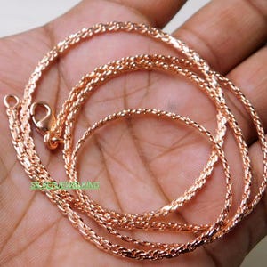 Könnte beinhalten: Eine Halskette aus Roségold mit einem Karabinerverschluss. Die Kette besteht aus kleinen, aufwendig verbundenen Segmenten, die ein strukturiertes Aussehen erzeugen. Die Halskette wird auf einer Hand präsentiert, um ihre Farbe und ihr Design zu zeigen.