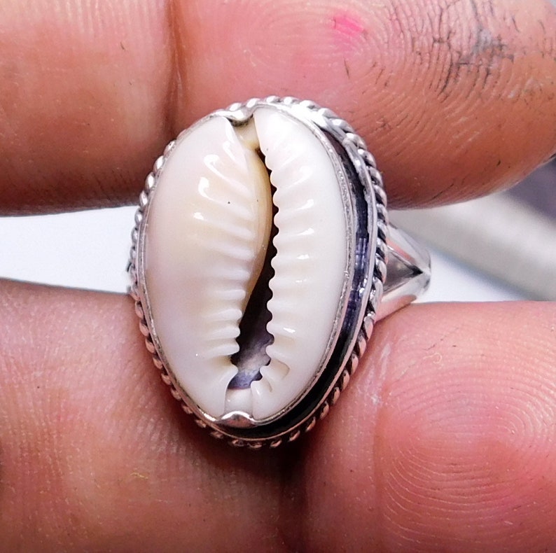 Op de afbeelding: Een zilveren ring met een witte kauri schelp in het midden gezet. De ring heeft een touwachtig ontwerp rond de rand van de zetting.