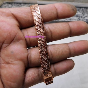 Może przedstawiać: Bransoletka w kolorze miedzi o teksturowanym, tkanym wzorze. Bransoletka jest prostokątna i posiada zapięcie. Obraz jest zbliżeniem, ukazującym skomplikowane detale metaloplastyki. Bransoletka jest trzymana w dłoni.