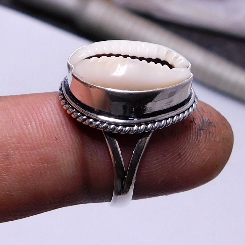 Op de afbeelding: Een zilveren ring met een witte kauri schelp in het midden gezet. De ring heeft een touwachtig ontwerp rond de rand van de zetting.