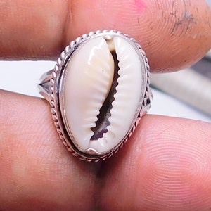 Op de afbeelding: Een zilveren ring met een witte kauri schelp in het midden. De ring heeft een touwachtig ontwerp rond de rand.