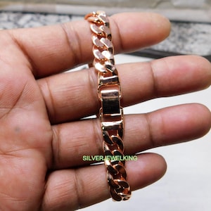 Może przedstawiać: Bransoletka z łańcuchem typu curb ze złota różowego z bezpiecznym zapięciem. Bransoletka posiada przeplatające się ogniwa, tworząc odważny dodatek. Tekst "SILVERJEWELKING" jest widoczny na bransoletce.