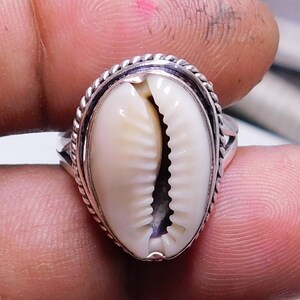 Op de afbeelding: Een zilveren ring met een witte kauri schelp in het midden gezet. De ring heeft een touwachtig ontwerp rond de rand van de zetting.