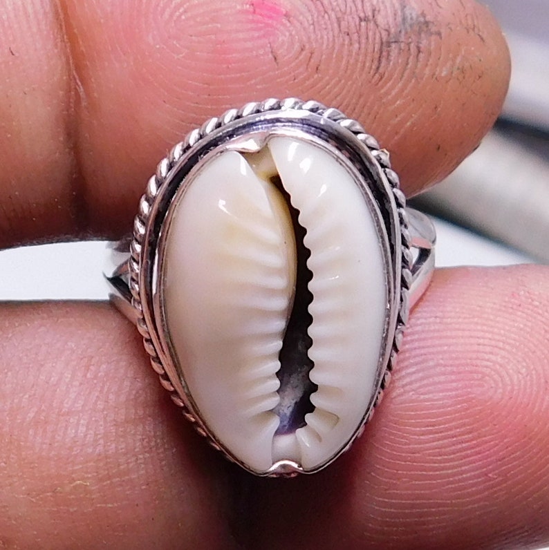 Op de afbeelding: Een zilveren ring met een witte kauri schelp in het midden gezet. De ring heeft een touwachtig ontwerp rond de rand van de zetting.