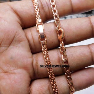 May include: A rose gold-toned chain bracelet featuring a woven wheat link design on one side and a flat curb link design on the other, connected by a rectangular clasp and a lobster claw clasp, with the text SILVERJEWELKING printed on the metal.