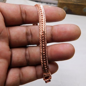 Puede incluir: Una pulsera de color cobre con un diseño de cadena de espiga. La pulsera está hecha de piezas de metal unidas y tiene un cierre seguro. El tono cálido, rojizo-marrón del metal le da a la pulsera un aspecto clásico.