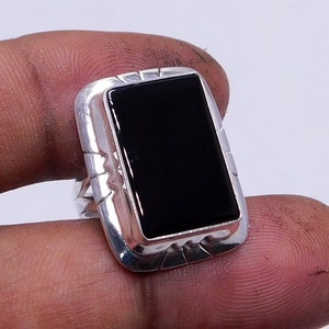 Pode incluir: Um anel retangular de ônix preto com uma banda de prata. A pedra de ônix é colocada em uma moldura retangular de prata com bordas decorativas. O anel é mostrado em um fundo neutro.