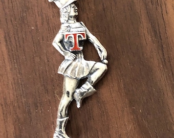Op de afbeelding: Zilveren bedel met een majorette van een marchingband in een dynamische pose. De figuur draagt een hoed, rok en laarzen, met een rode "T" op de borst. De bedel is opgehangen aan een kleine lus.