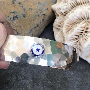 May include: Silver-toned cuff bracelet with a hammered texture. The bracelet features a circular emblem with the text "American Legion Auxiliary" and a blue star on a white background. The bracelet is held by a hand.