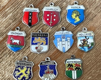 May include: A collection of ten vintage silver-toned shield charms, each with a different colorful enamel design. The charms feature city names like Amsterdam, Haarlem, and Rotterdam. They are likely souvenir or travel-themed jewelry.