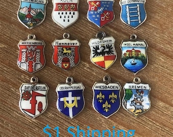 May include: A collection of twelve silver-toned shield-shaped charms, each with a different colorful enamel design. The charms feature various city names, including Garmisch-Partenkirchen, Köln, and Bremen. The charms are displayed on a wooden surface.