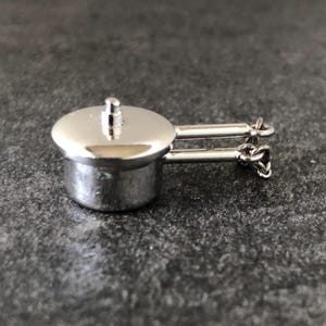 Puede incluir: Una pequeña olla plateada brillante con tapa y dos asas. La pequeña olla es probablemente un amuleto o colgante, con un diseño detallado y un acabado pulido. Las asas están unidas con pequeños bucles.