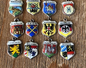 May include: A collection of vintage shield-shaped charms with various city names and emblems. Each charm is silver-toned with colorful enamel details, including images of landmarks, coats of arms, and historical figures. The charms are displayed on a wooden surface.